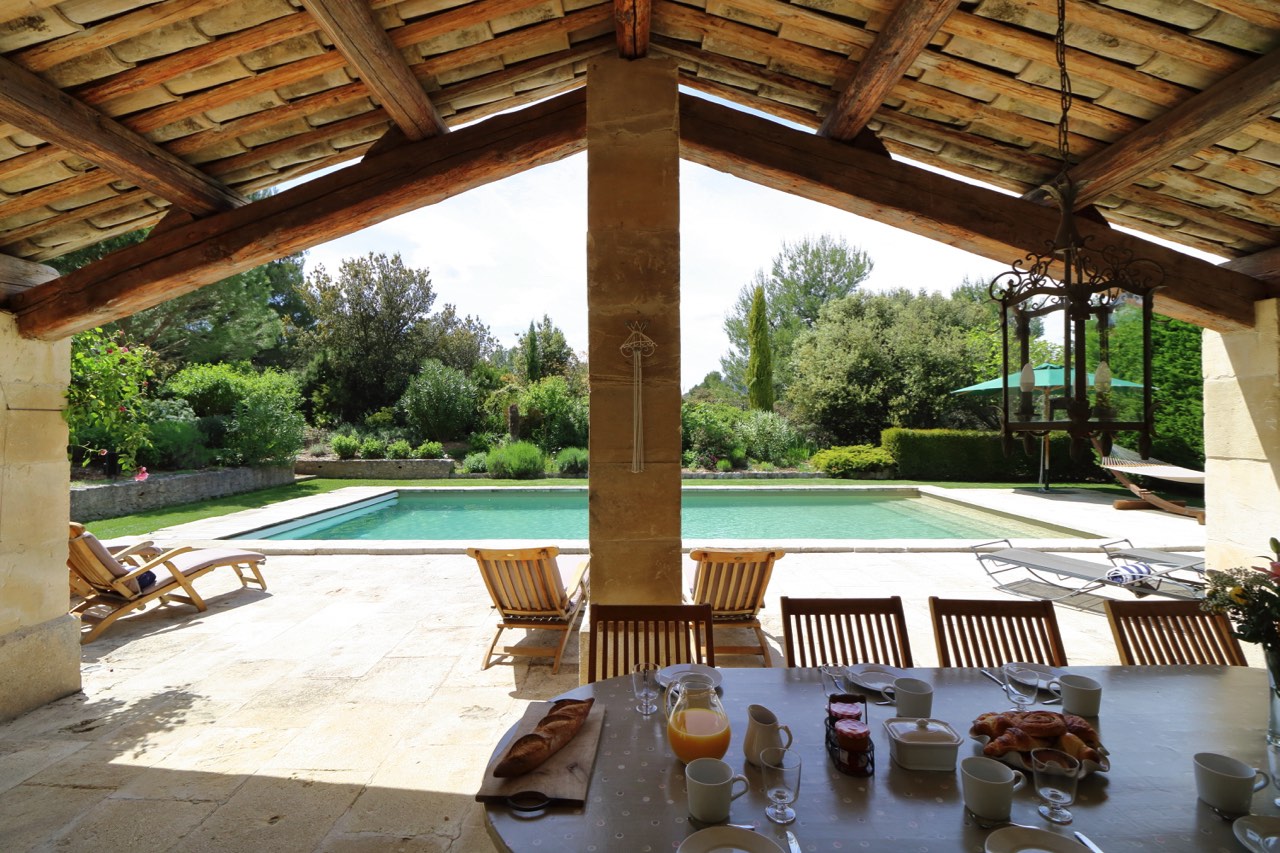 Outdoor breakfast setup under a covered patio with a view of a swimming pool and lush garden, featuring a table with croissants, juice, and coffee.